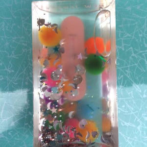 Op de afbeelding: Een heldere, rechthoekige, met vloeistof gevulde container met kleurrijke pompons, pailletten en een roze object erin. De container heeft een doorzichtige lus aan de bovenkant. De pompons zijn oranje, groen, geel en blauw. De pailletten zijn zilverkleurig en stervormig.