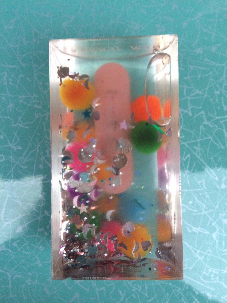 Op de afbeelding: Een heldere, rechthoekige, met vloeistof gevulde container met kleurrijke pompons, pailletten en een roze object erin. De container heeft een doorzichtige lus aan de bovenkant. De pompons zijn oranje, groen, geel en blauw. De pailletten zijn zilverkleurig en stervormig.