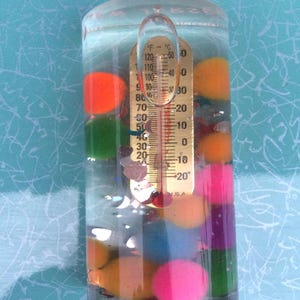 Op de afbeelding: Een heldere, cilindrische vloeistofthermometer met een goudkleurige temperatuurmeter. De thermometer is gevuld met water en kleurrijke bollen. De temperatuurmeter geeft Fahrenheit en Celsius aan. De bollen zijn oranje, groen, roze, paars en blauw.