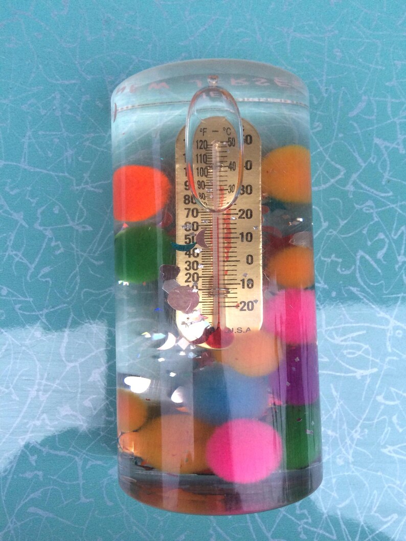 Op de afbeelding: Een heldere, cilindrische vloeistofthermometer met een goudkleurige temperatuurmeter. De thermometer is gevuld met water en kleurrijke bollen. De temperatuurmeter geeft Fahrenheit en Celsius aan. De bollen zijn oranje, groen, roze, paars en blauw.