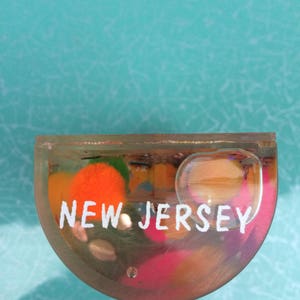 Op de afbeelding: Een heldere, halfronde presse-papier met de tekst "NEW JERSEY" in het wit. De presse-papier bevat kleurrijke, abstracte vormen in oranje, roze en groen. De achtergrond is lichtblauwgroen.