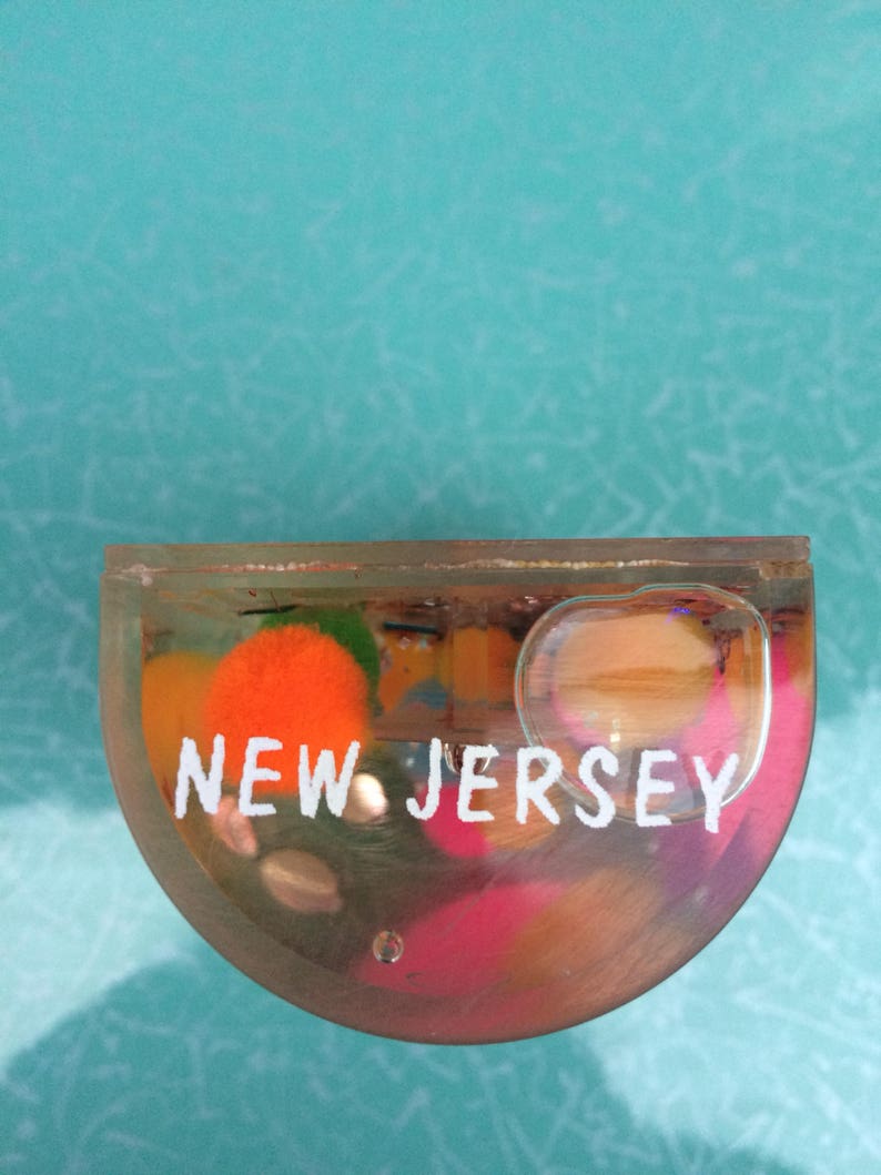 Op de afbeelding: Een heldere, halfronde presse-papier met de tekst "NEW JERSEY" in het wit. De presse-papier bevat kleurrijke, abstracte vormen in oranje, roze en groen. De achtergrond is lichtblauwgroen.