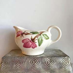 May include: A small, white ceramic creamer with a handle and spout. The creamer is decorated with pink flowers, green leaves, and brown stems. The creamer is sitting on a silver, textured surface.
