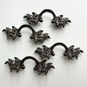 May include: A set of four dark bronze-toned metal drawer pulls. Each pull features a central arched handle flanked by ornate, symmetrical scrollwork and leaf-like detailing on both sides, creating a cohesive look for furniture hardware.