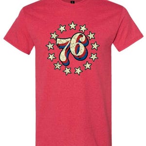 Può includere: T-shirt rosso con un design retrò. Il numero "76" è in rosso, bianco e blu, circondato da stelle. La maglietta ha un girocollo e maniche corte.