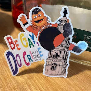 May include: A sticker featuring a cartoon character with orange fur and a black and orange jersey. The character is holding a building with a clock tower. The sticker has the text "Be Gay, Do Crime" in rainbow colors.