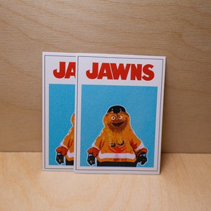 A photo of 2 (two) stickers standing against a light wood grain wall that look like the famous JAWS movie poster but feature a cartoon image of NHL Philadelphia Flyers mascot Gritty.