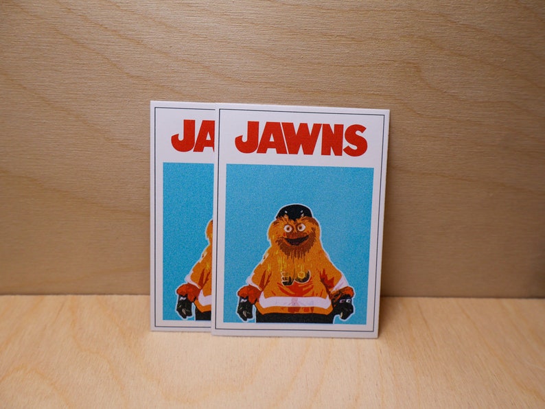 A photo of 2 (two) stickers standing against a light wood grain wall that look like the famous JAWS movie poster but feature a cartoon image of NHL Philadelphia Flyers mascot Gritty.