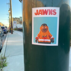 photo of a green street pole with a rectangular sticker of NHL Philadelphia Flyers Mascot Gritty, in the style of the JAWS movie poster, that reads JAWNS instead of JAWS.