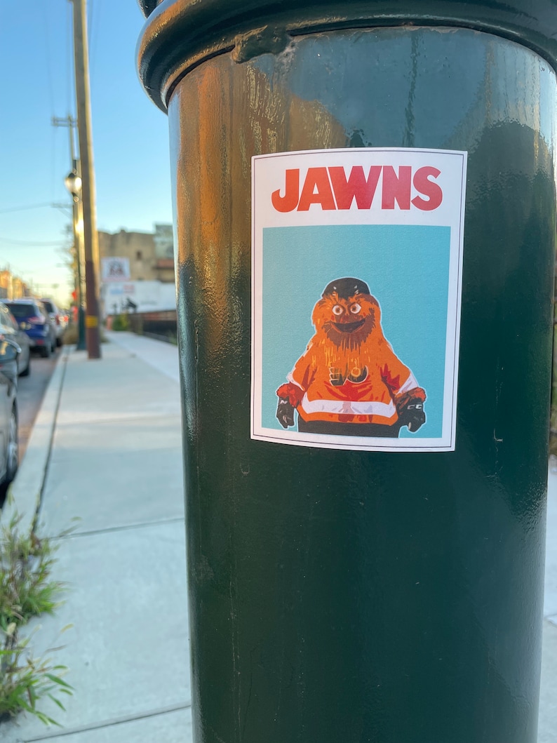 photo of a green street pole with a rectangular sticker of NHL Philadelphia Flyers Mascot Gritty, in the style of the JAWS movie poster, that reads JAWNS instead of JAWS.