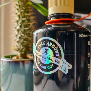 May include: Black water bottle with a holographic sticker that says "F**k Around Philadelphia, PA and Find Out". The sticker features a Liberty Bell graphic. The bottle has a beige lid and an orange carrying strap.