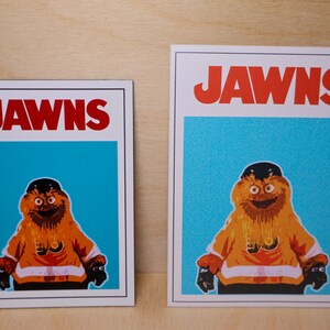 Close-up photo of a pair of sticker and magnet with the same graphic on them. The graphic looks like the JAWS movie poster but features a cartoon image of NHL Philadelphia Flyers mascot Gritty.