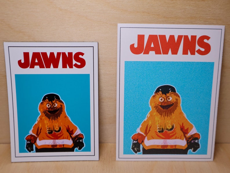 Close-up photo of a pair of sticker and magnet with the same graphic on them. The graphic looks like the JAWS movie poster but features a cartoon image of NHL Philadelphia Flyers mascot Gritty.