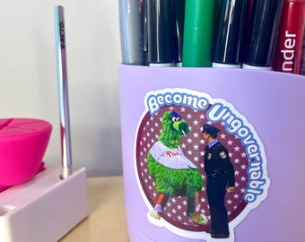 May include: A purple pencil holder with a sticker on the front. The sticker features a green cartoon character with a baseball cap and a police officer. The sticker says "Become Ungovernable".