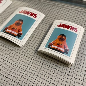 Photo of 3 (three) stacks of stickers that have Gritty on them, sitting on top of a surface with a tight grid and measurements.The stickers look like the JAWS movie poster.