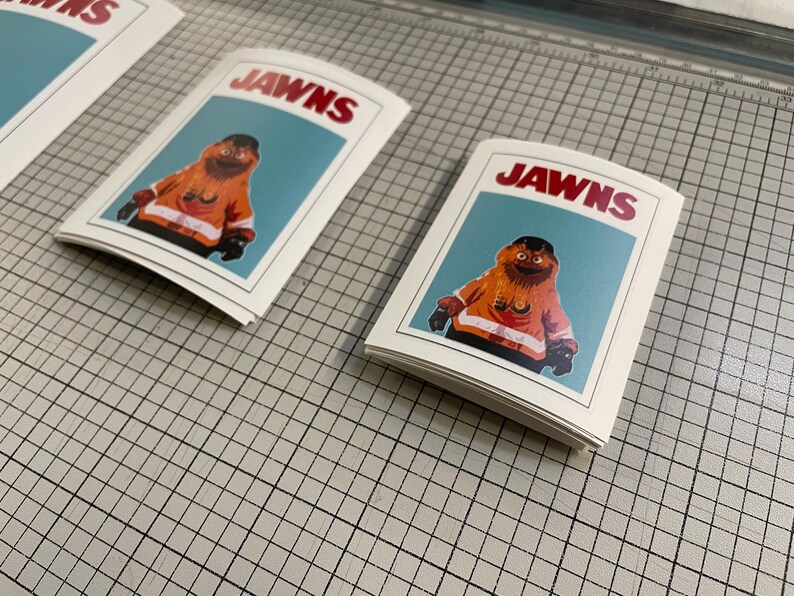 Photo of 3 (three) stacks of stickers that have Gritty on them, sitting on top of a surface with a tight grid and measurements.The stickers look like the JAWS movie poster.