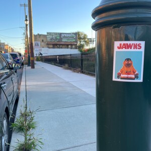 Landscape oriented photo of a sidewalk with a green pole with a rectangular sticker of NHL Philadelphia Flyers Mascot Gritty, in the style of the JAWS movie poster, that reads JAWNS instead of JAWS.