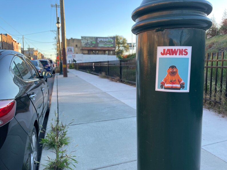 Landscape oriented photo of a sidewalk with a green pole with a rectangular sticker of NHL Philadelphia Flyers Mascot Gritty, in the style of the JAWS movie poster, that reads JAWNS instead of JAWS.