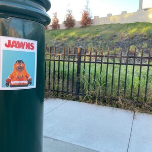 photo of a green street pole with a rectangular sticker of NHL Philadelphia Flyers Mascot Gritty, in the style of the JAWS movie poster, that reads JAWNS instead of JAWS. There is a grassy hillside/freeway beyond the pole.