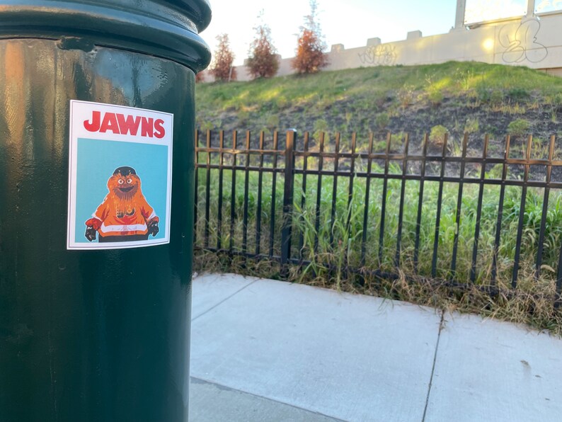 photo of a green street pole with a rectangular sticker of NHL Philadelphia Flyers Mascot Gritty, in the style of the JAWS movie poster, that reads JAWNS instead of JAWS. There is a grassy hillside/freeway beyond the pole.