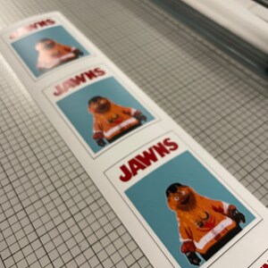 Photo of a vertical strip of stickers that have Gritty on them, sitting on top of a surface with a tight grid and measurements.The stickers look like the JAWS movie poster.