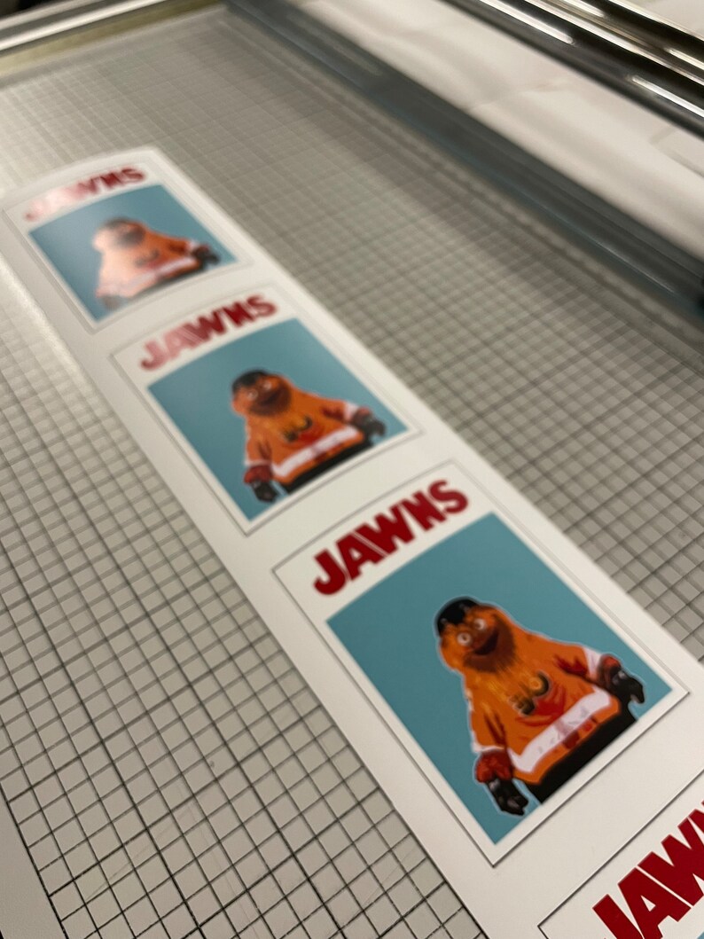 Photo of a vertical strip of stickers that have Gritty on them, sitting on top of a surface with a tight grid and measurements.The stickers look like the JAWS movie poster.