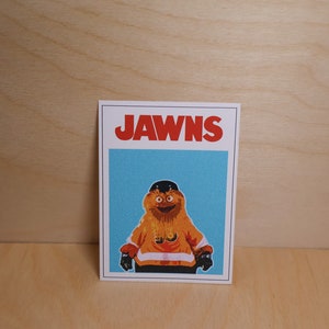 A photo of a rectangular sticker standing against a light wood grain wall that looks like the famous JAWS movie poster but features a cartoon image of NHL Philadelphia Flyers mascot Gritty.