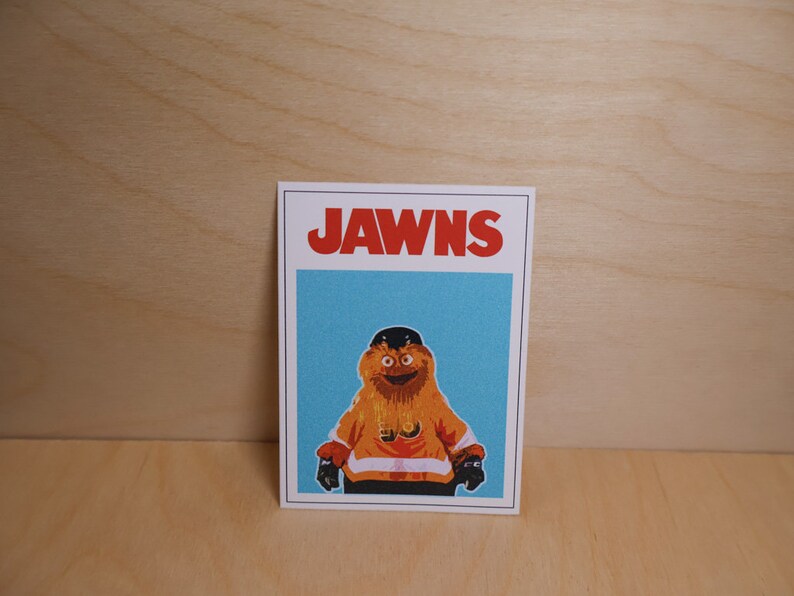 A photo of a rectangular sticker standing against a light wood grain wall that looks like the famous JAWS movie poster but features a cartoon image of NHL Philadelphia Flyers mascot Gritty.