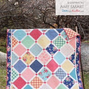 May include: A quilt with a lattice pattern in shades of pink, blue, yellow, and green. The quilt is made of various fabrics with floral, polka dot, and plaid patterns. The quilt is hanging on a clothesline in front of a tree with white blossoms.
