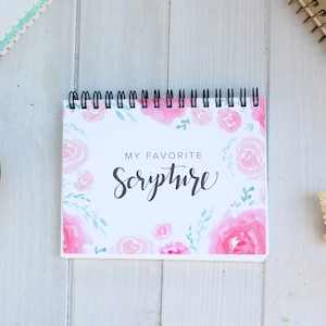 May include: A white spiral bound notebook with a pink and green floral design. The notebook says "My Favorite Scripture" in black script.