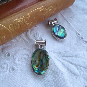 May include: Three oval pendants featuring iridescent abalone shells set in silver. The pendants are displayed on a vintage book and white lace, highlighting their natural blue, green, and gold hues.