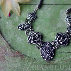 Può includere: Una collana color argento dal design ornato. La collana presenta pendenti quadrati e ovali con una texture a pois e motivi a spirale. La catena è in stile intrecciato. Lo sfondo è una superficie verde con un fiore.