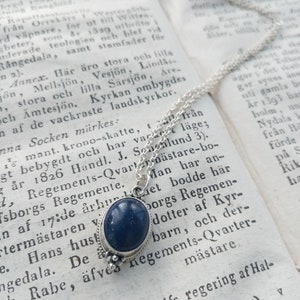 May include: A silver chain necklace with a dark blue gemstone pendant. The pendant is oval-shaped and has a silver setting. The necklace is lying on a page of a book with text in a foreign language.