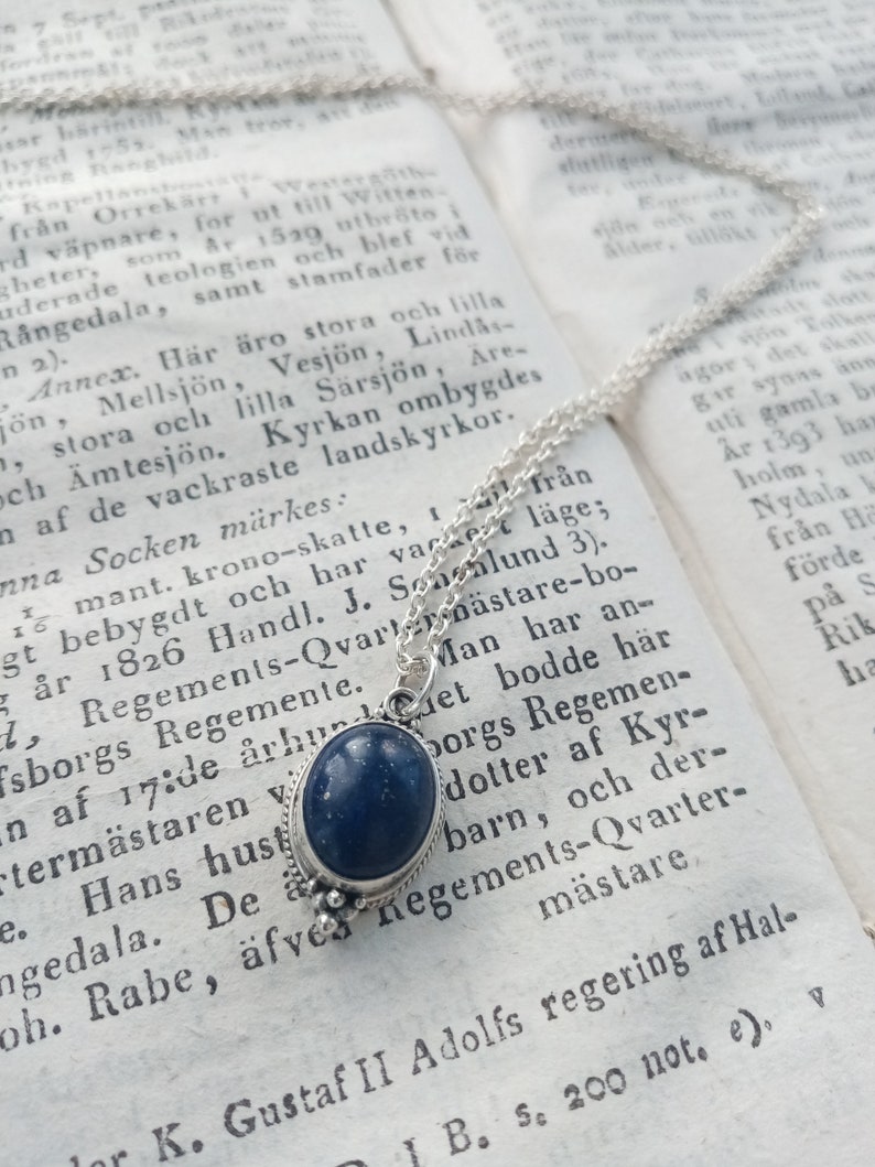 May include: A silver chain necklace with a dark blue gemstone pendant. The pendant is oval-shaped and has a silver setting. The necklace is lying on a page of a book with text in a foreign language.