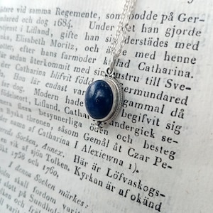 May include: A silver necklace with a blue lapis lazuli stone pendant. The pendant is set in a silver bezel and hangs from a delicate silver chain. The necklace is displayed on a page of an old book with text in Swedish.