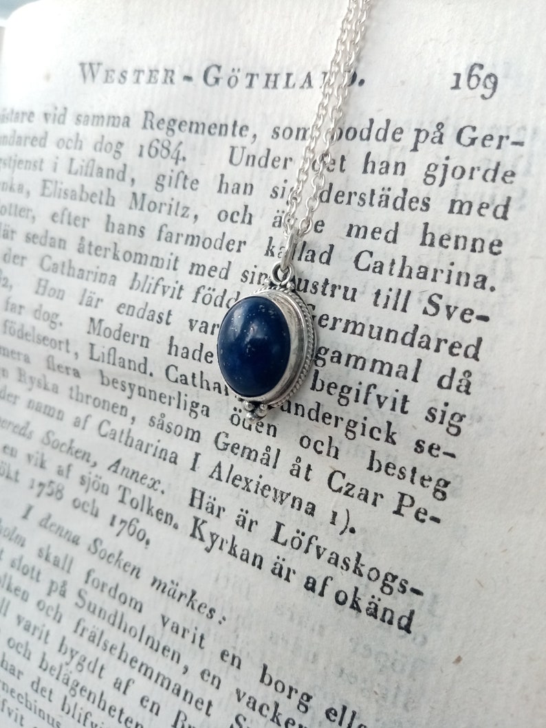 May include: A silver necklace with a blue lapis lazuli stone pendant. The pendant is set in a silver bezel and hangs from a delicate silver chain. The necklace is displayed on a page of an old book with text in Swedish.