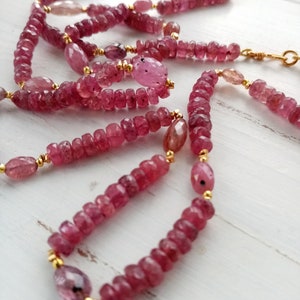 May include: A long pink gemstone necklace with gold accents. The necklace is made of small, faceted pink gemstones and gold beads. The necklace is layered and has a delicate, feminine look.