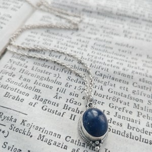 May include: A silver chain necklace with a blue gemstone pendant. The pendant is oval-shaped and has a silver bezel setting. The chain is delicate and has a simple design.