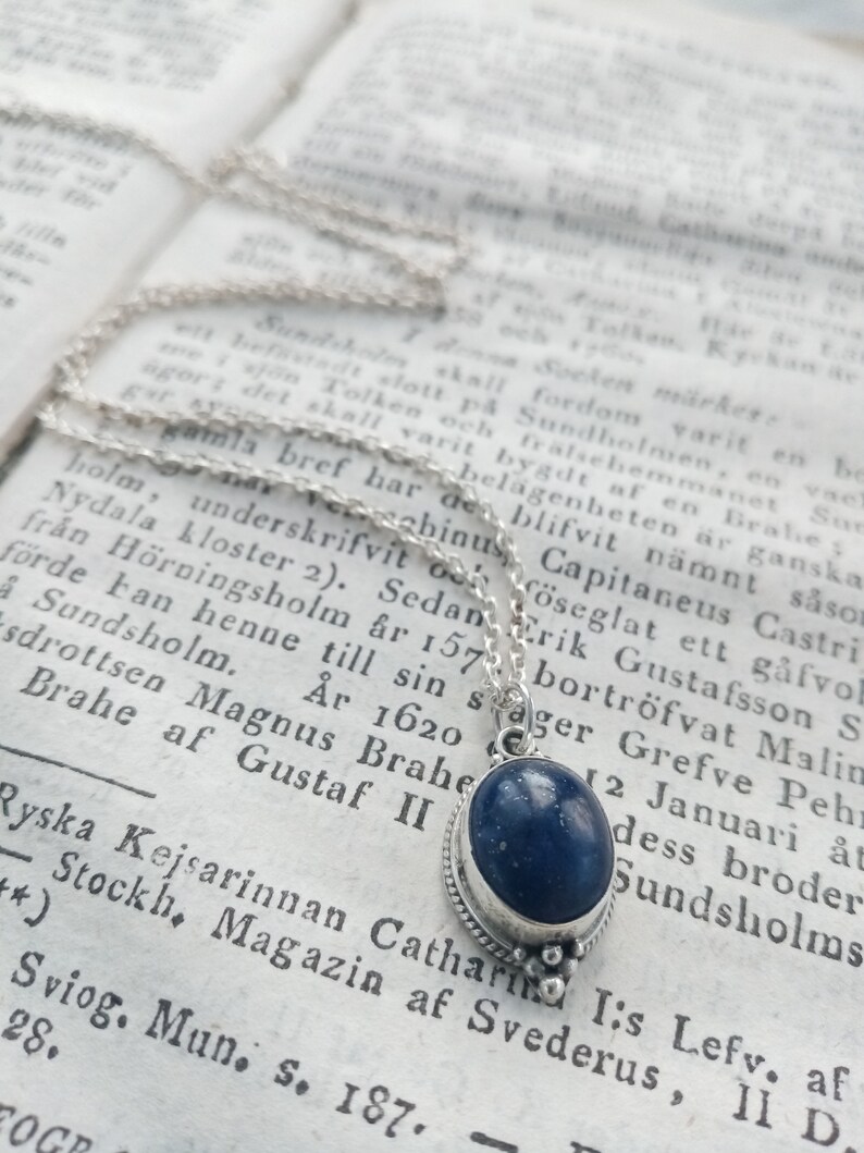 May include: A silver chain necklace with a blue gemstone pendant. The pendant is oval-shaped and has a silver bezel setting. The chain is delicate and has a simple design.