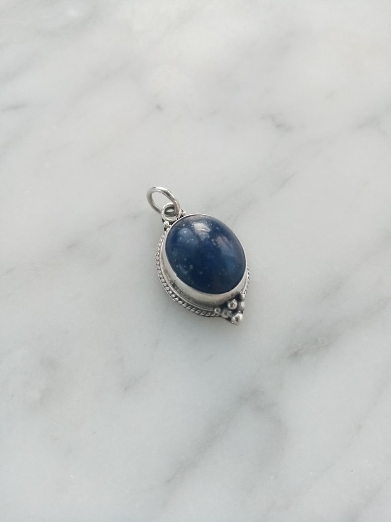 May include: A silver pendant with a dark blue oval gemstone set in a silver bezel. The pendant has a silver bail for attaching to a chain.