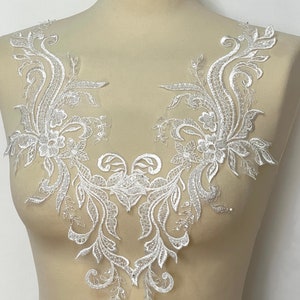 May include: White lace applique with a floral design. The applique is shaped like a V and has a delicate, intricate pattern.