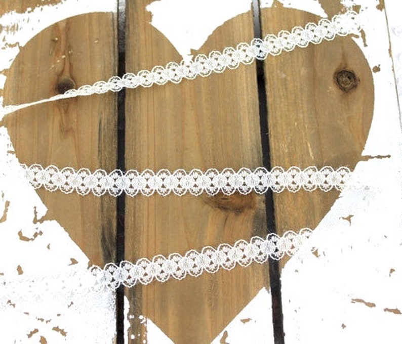 May include: White lace trim with a delicate floral pattern. The lace is shown on a wooden heart background.