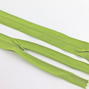 Pode incluir: Duas fitas de zíper verdes com um cursor de zíper em uma extremidade. As fitas de zíper estão dispostas sobre uma superfície branca.