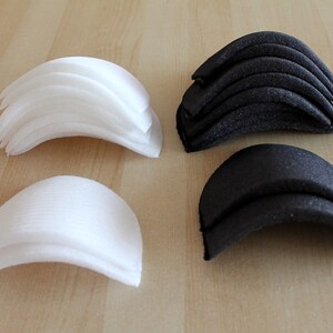 Può includere: Due set di imbottiture per reggiseno in tessuto, uno bianco e uno nero. Ogni set è impilato in una forma curva.