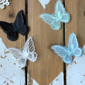 Op de afbeelding: Vier vlindervormige geborduurde patches met vleugels van transparante organza en gedetailleerd stiksel. De set bevat een zwarte, een witte, een licht mintgroene en een lichtblauwe vlinderpatch, elk met een verfijnde geschulpte rand.