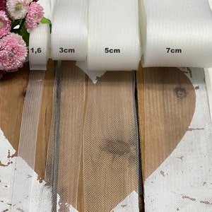 Könnte beinhalten: Vier Rollen weißer Tüllstoff, jede mit einer anderen Breite: 1,6 Zoll, 3 cm, 5 cm und 7 cm. Der Stoff liegt auf einer Holzoberfläche.