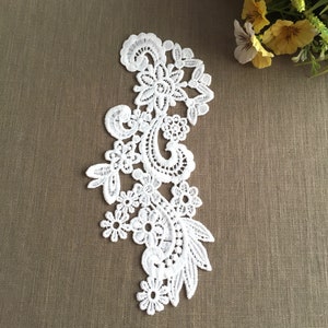 Op de afbeelding: Witte kant applique met een bloemenmotief. De kant is delicaat en ingewikkeld, met een verscheidenheid aan bloemen en bladeren. De applique is perfect om een ​​toets van elegantie aan elk project toe te voegen.