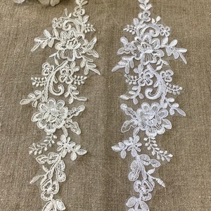 Puede incluir: Dos apliques de encaje blanco con diseños florales. Los apliques están sobre un fondo de tela beige.