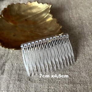 May include: A clear plastic hair comb with a row of teeth. The comb is 7cm long and 4.5cm wide. The comb is on a textured surface with a gold leaf-shaped dish.