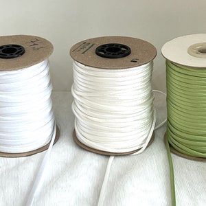 Könnte beinhalten: Drei Spulen mit weißem und grünem Satinband. Die Spulen sind aus Pappe und haben einen Plastikkern.